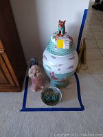 Full view of floor vase with lid, two ceramic figurines, decorative bowl with balls, all placed on carpet near wooden cabinet.