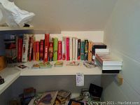 Shelf containing various books mostly about playing cards, card games, and magic. Several boxed sets of playing cards and small children's books or games. Items arranged in a row on a white shelf.