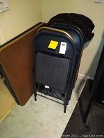 Photos of the lot showing one rectangular folding card table with brown surface and black edge, accompanied by four black metal folding chairs with padded gray striped seats. Items are stacked and placed against a wall on a carpeted floor.