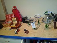 Front view of lot including 4 margarita glasses, rocking chair, sewing basket, red suitcase and dog figurine on table.