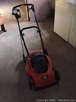 Black & Decker orange and black electric lawn mower, shown from front angle, with visible handle and wheels on concrete floor indoors.
