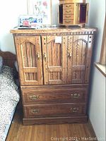 Full view of wooden tall boy dresser showing 2 doors and 2 drawers, brass hardware, and top surface items including jewelry box and framed photo.