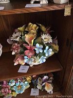 Porcelain flower arrangement on wooden shelf, showing multiple flowers in yellow, pink, white, and blue colors.