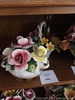 Porcelain flower arrangement with pink and cream roses and yellow flowers, placed on wooden shelving