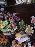 Two porcelain floral arrangements by Capodimonte displayed on a glass shelf and wooden table, showing vibrant flowers and detailed leaves.