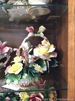 Front and side views of two Capodimonte porcelain floral arrangements with yellow and pink flowers, green foliage, and basket handles with bow accents.