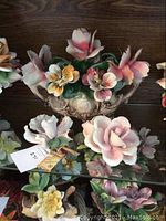 Photo showing three Capodimonte floral porcelain pieces on a glass shelf with detailed multi-colored flowers on an ornate base and two smaller flower sculptures.