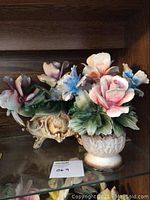 Front view of one flower arrangement featuring pink and blue flowers with green leaves on a cream decorative base displayed on a glass shelf.