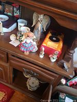 Photo showing glass angel with light-up base and other angel figurines, porcelain doll, teacup and saucer, and various small decor items arranged on wooden surface.
