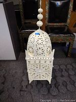 Front view of white Moroccan hand-cut iron lantern on carpet with two dark decorative chairs with studs and colored insets in background.