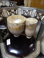 Full view of the pair of Moroccan drums placed on a decorative chair with intricate metal and leather work. The drums show natural camel rawhide surfaces and ceramic bases, with rope bindings at the base of the drum heads.