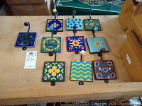 Overview of ten colorful hand painted ceramic tile wall hooks arranged on table.