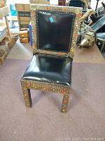 Front view of single Moroccan chair showing wood frame with metal overlay, black leather seat and back with nailhead trim and colored ornamental accents.