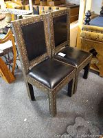 Two Moroccan side chairs placed side by side showing full front and side views, black leather upholstery and metal inlay