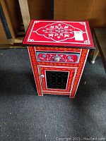 Front view of rectangular Moroccan style wooden side table painted with vibrant red, blue, yellow, white, and black traditional motifs. One drawer and one cabinet door with ceramic knobs visible.