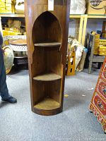 Wooden corner shelving unit with three shelves and Moroccan style arch-shaped cut-out front, standing on floor with surrounding items.