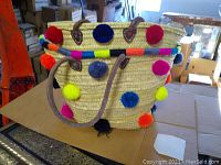 Wide view of one basket showing straw weaving, leather handles, and colorful pom poms around the exterior.