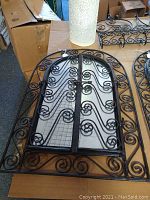 Photo showing two wrought iron framed arched mirrors with decorative scrollwork lying flat on cardboard.