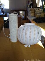 Two white Moroccan tin metal lamps on a table, one tall cylindrical lamp with floral perforations and one round ribbed lamp shade with vertical perforated stripes.