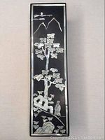 Full front view of rectangular black trinket box with mother of pearl inlaid decorations including trees, rocks, figure, and mountains.