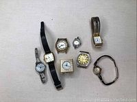 Seven vintage wristwatches and miniature travel alarm clocks arranged on beige surface, showing variety of watch faces and bands.