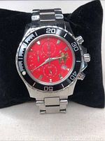 Front view of wristwatch showing red dial with gold RCMP mounted officer emblem, black bezel with minute markings, metal case and band.