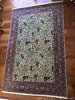 Full view of rectangular rug laid on hardwood floor showing ivory field with hunting figures and burgundy border