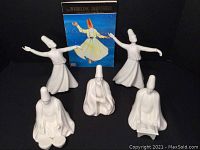 Set of five white ceramic Turkish Whirling Dervishes figurines and an illustrated book, arranged on a black surface.