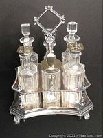 Front angle view of vintage cruet set with clear glass bottles in a silver plated holder on decorative feet, showing stoppers and pierced lids.