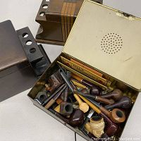 Metal humidor box open showing several vintage smoking pipes inside with a few paper labels or instructions.