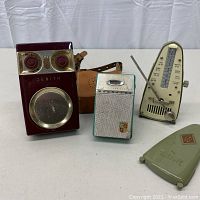 Front view of Zenith Royal 500 transistor radio, Magnavox transistor radio, Taktell metronome, and green plastic metronome cover arranged on table.