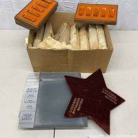 Photo showing two wooden watch display trays with padded orange slots on top of a cardboard box containing wrapped items; clear acrylic Bulova Watches display holders and a burgundy velvet star-shaped Longines Wittnauer display piece in foreground.