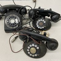 Three vintage black rotary dial telephones tangled with their cords on a white surface.
