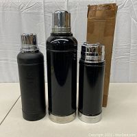 Three black Stanley thermoses with chrome tops and bases, standing upright, with original cardboard box behind