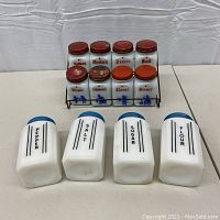 Front view of the full lot showing the metal spice rack with eight small milk glass spice jars with red lids, and in front four larger milk glass canisters with blue lids labeled with kitchen staples.