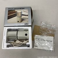 Overview photo showing the Braun HL-70 personal fan, power cord, instruction booklet, plastic mounting brackets, all housed inside the original Styrofoam insert and box with Braun branding.