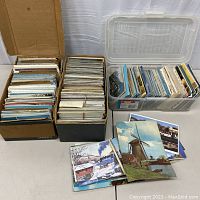Two cardboard boxes and one plastic container filled with stacks of postcards on a white table with loose cards showing scenic railroad images