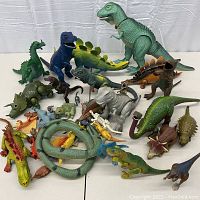 Lot of vintage dinosaur, dragon, and reptile figures arranged on a white surface, showing various colors and sizes, some articulated limbs visible.