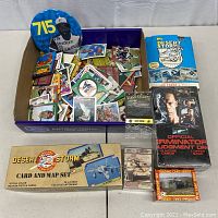 Photo of a box with assorted vintage baseball and football trading cards, Desert Storm related sets, and a Terminator 2 card pack, plus collectible pin-back button.