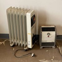 Wide view showing both heaters side by side against a wall on a brown floor; DeLonghi is oil-filled radiator style, Lasko is ceramic fan type.