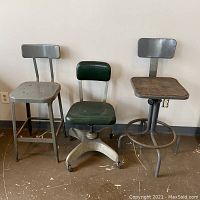 Three seating pieces side by side against wall: perforated metal chair, green vinyl swivel chair, adjustable shop stool
