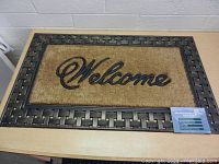 Large rectangular welcome mat with natural fiber center displaying 'Welcome' in black script and a sturdy black rubber frame with a woven pattern, lying on a table.