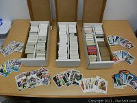 Three white storage boxes filled with NFL and MLB sports cards placed on a table with loose cards around them, showing fronts of various football and baseball players.