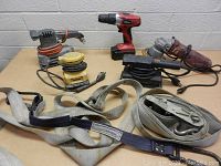 Photo showing assorted power tools including electric sanders, a cordless drill, and heavy-duty woven straps on a table.