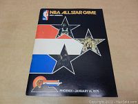 Front cover of the 1975 NBA All Star Game program showing the colorful red, white, and blue star cutouts with images of players and the event details. Held flat on a table.