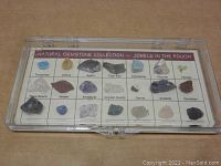 Top-down view of natural gemstone collection kit inside closed plastic case, showing 16 labeled gemstone specimens.