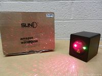Front and side view of black cube-shaped SunD laser light projector next to original Amazon warehouse packaging box with laser lights projected on floor.