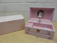 Pink rectangular musical jewelry box open showing interior compartments and spinning ballerina under a lid mirror with decorative ballerina and floral motifs on the outside.