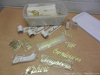 Top view of plastic container filled with gold-tone lettering words and rolls. Several words are laid out on the table: Mother, Grandpa, Husband, Nephew, Wife, Employees, Friends, and more.