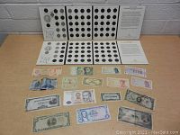 Top-down photo showing two open Fifty State Quarters booklets with empty slots arranged in rows, some quarters inserted, and many foreign banknotes spread out on the table surface below the booklets.